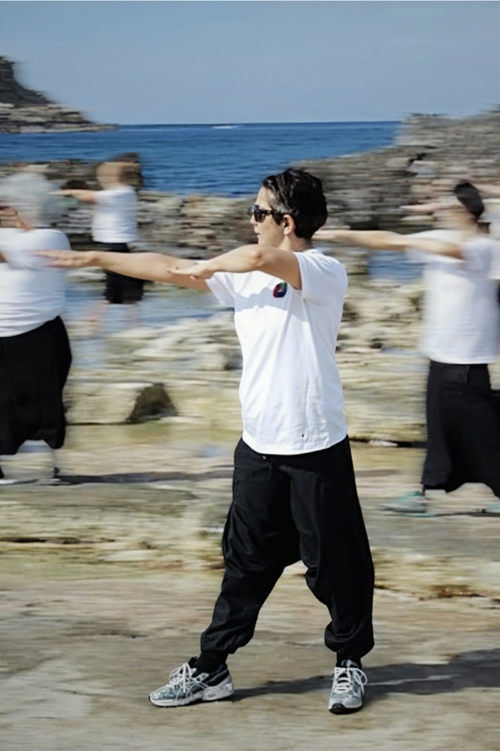 Group doing Tai Chi near the ocean, centered on woman in white shirt and black pants in motion, symbolizing flexible online healing.
