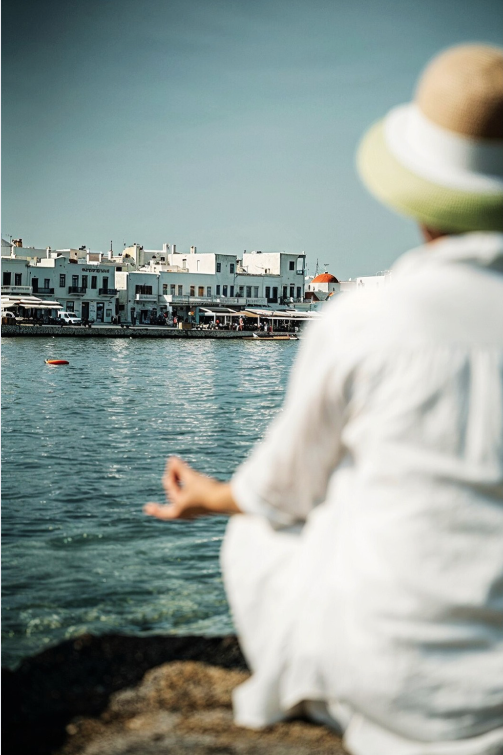 Person meditating by the sea in white clothing and sun hat, seen from behind, focusing on mindfulness and emotional healing.