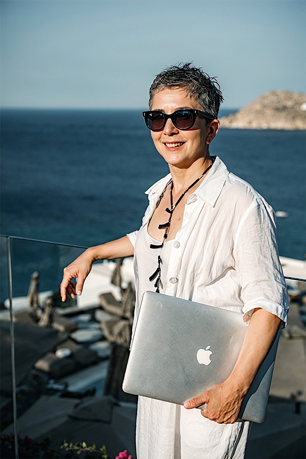 Smiling woman in white holding a laptop, standing on a balcony above the ocean, radiating confidence and transformation.